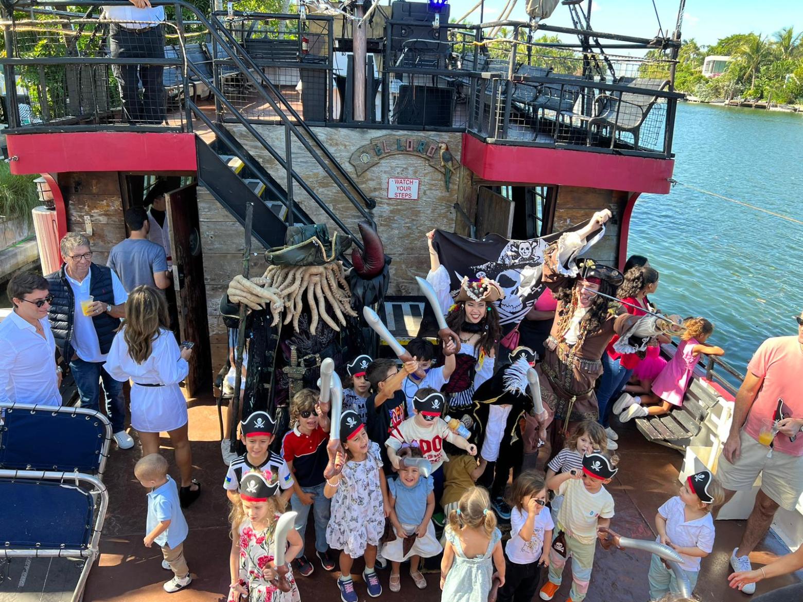 Group of kids with pirate hats and costumes on a ship with adults and a pirate flag.