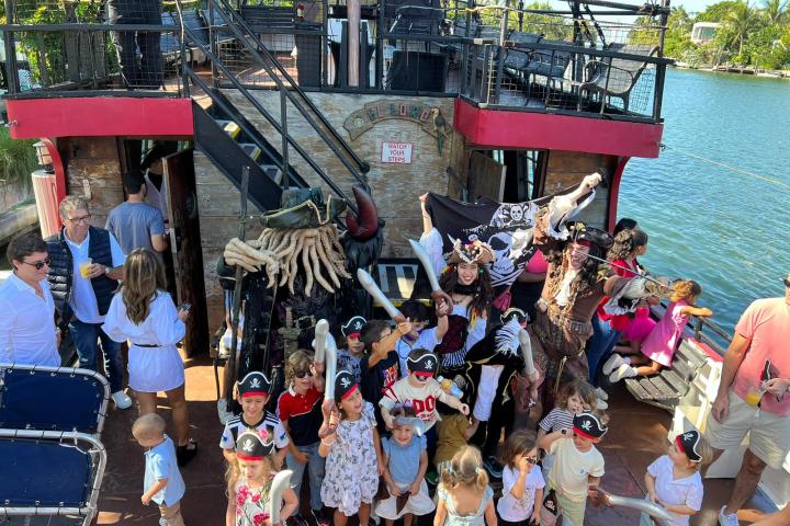 Group of kids with pirate hats and costumes on a ship with adults and a pirate flag.
