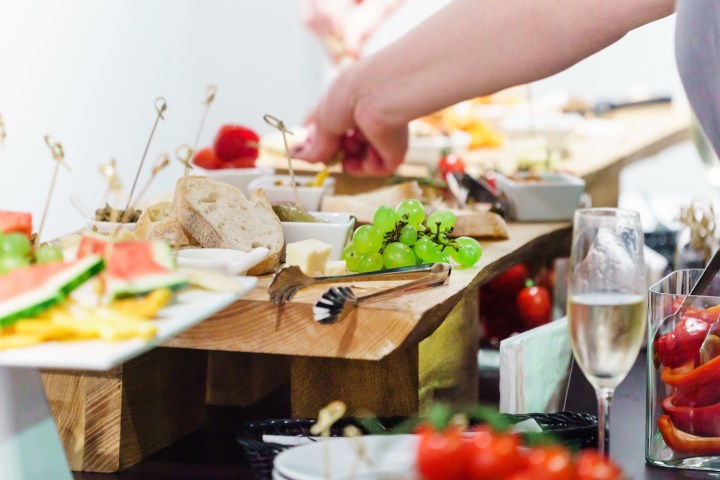 A buffet with bread, grapes, tomatoes, and champagne. A person reaches for food.