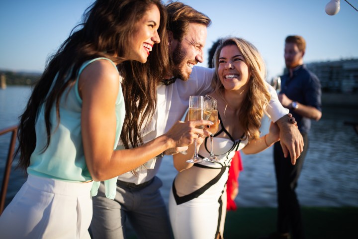 Three people laughing and holding champagne glasses at a sunny outdoor event by the water.