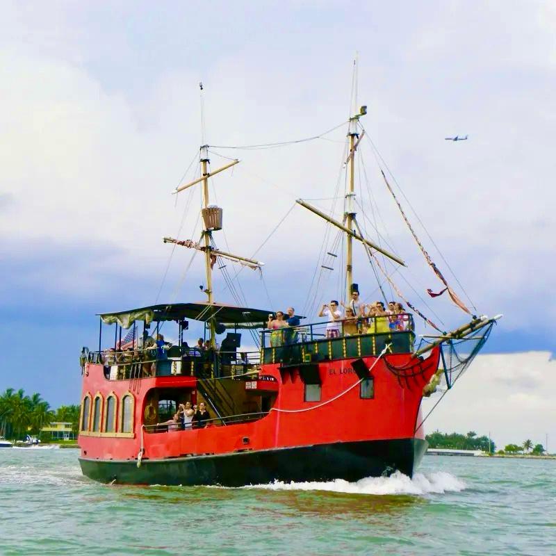 Red pirate-style ship with people aboard sailing near shore under a cloudy sky.