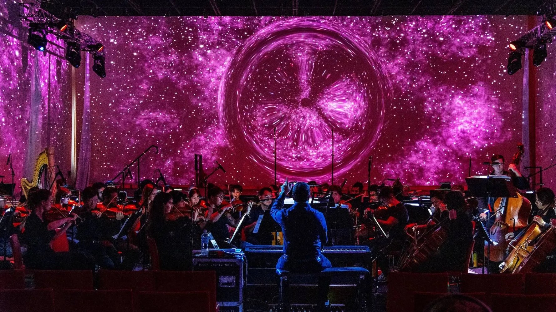Orchestra performing with conductor, pink galaxy visuals on screen in background.