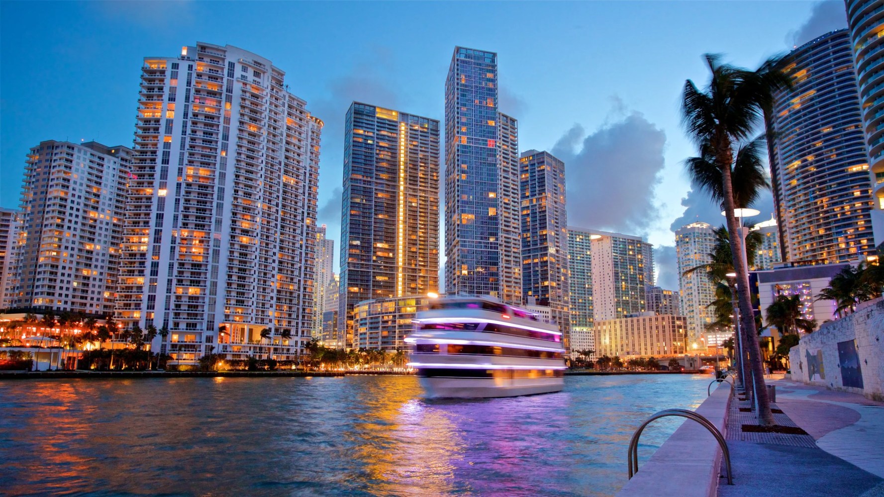 City skyline with tall buildings and a boat on the water at dusk.