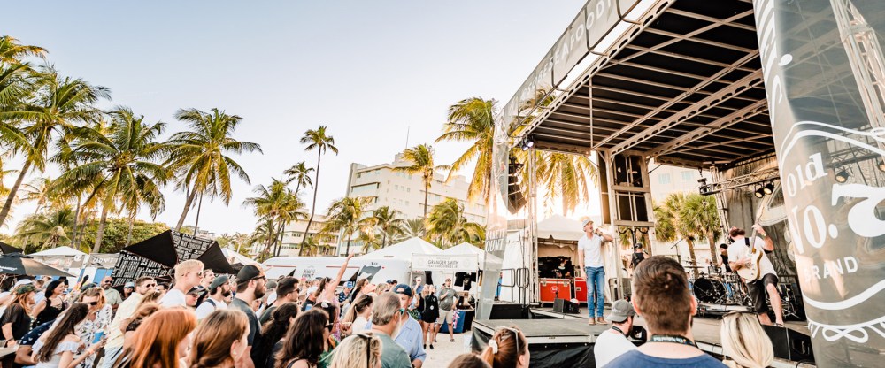 Outdoor concert with a crowd, palm trees, and a stage on a sunny day.