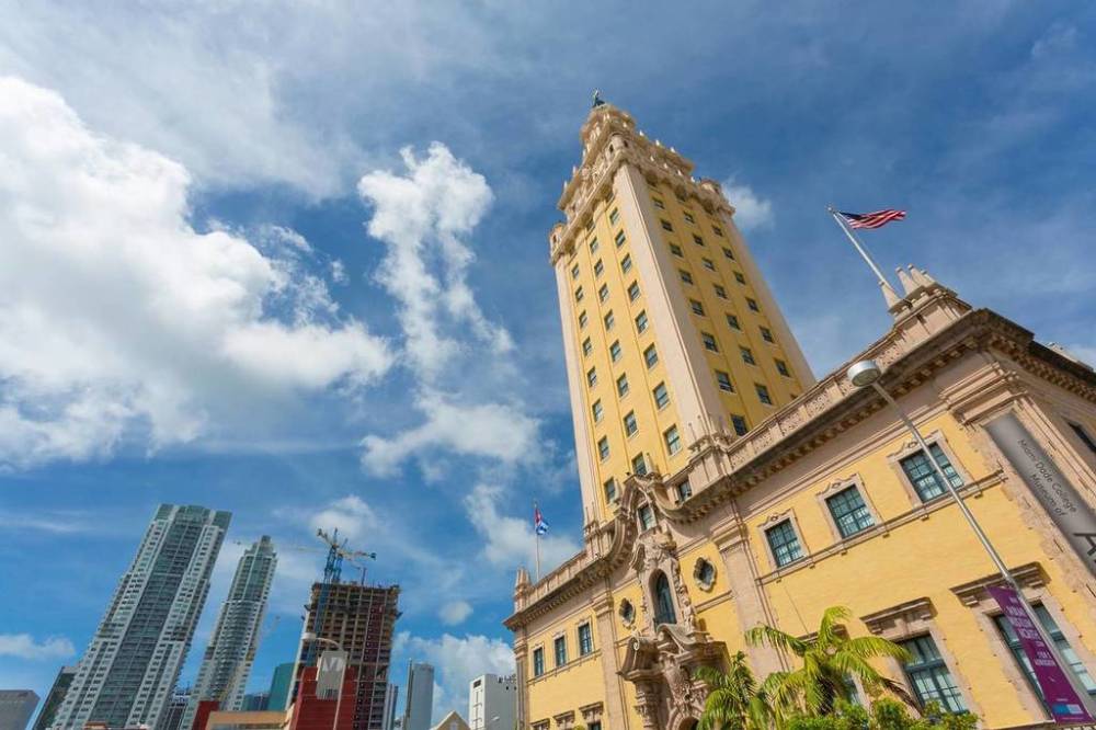 Tall yellow building with flag, skyscrapers and cloudy blue sky in background.