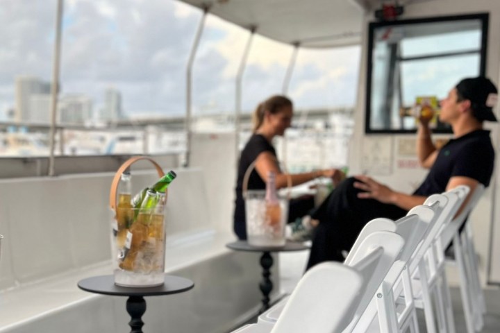 Two people on a boat with drinks on tables and cityscape blurred in the background.