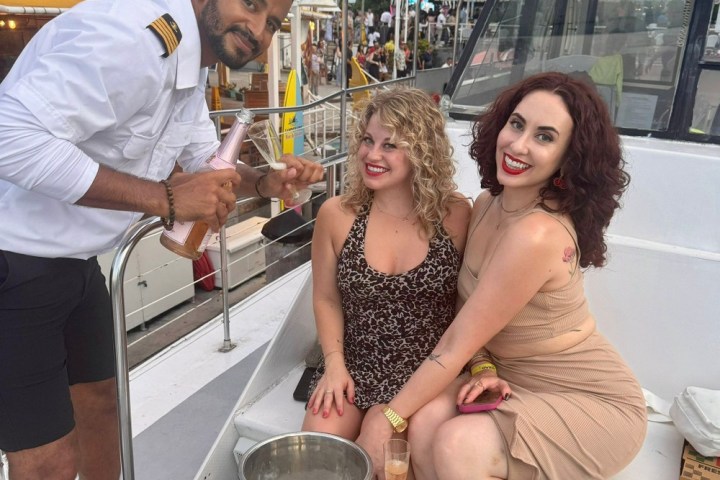Two women seated on a boat with a uniformed man pouring champagne nearby.