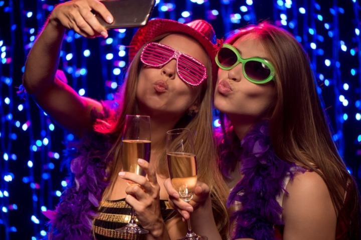 Two women in party attire taking a selfie with champagne, colorful glasses, and a bright backdrop.