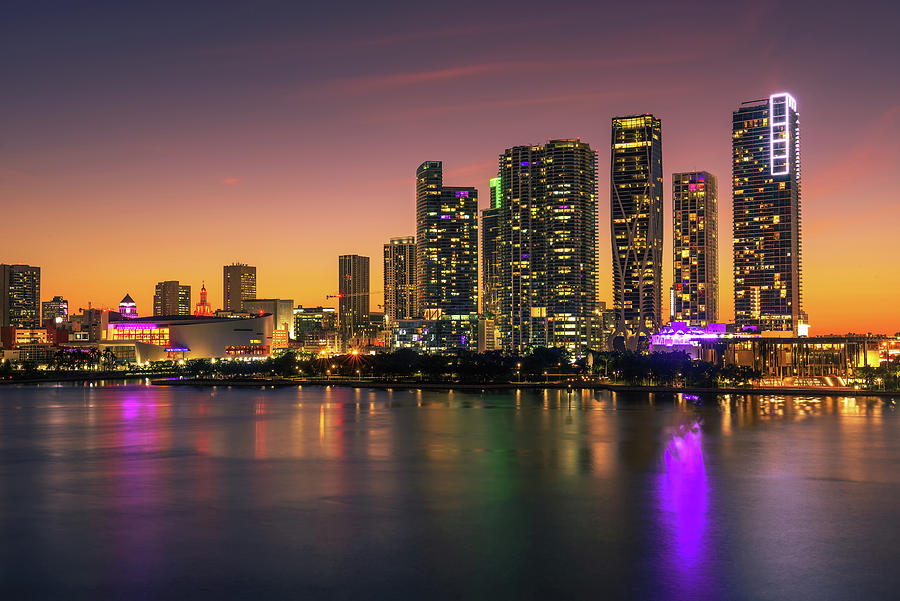 City skyline at sunset with illuminated skyscrapers reflecting on water.
