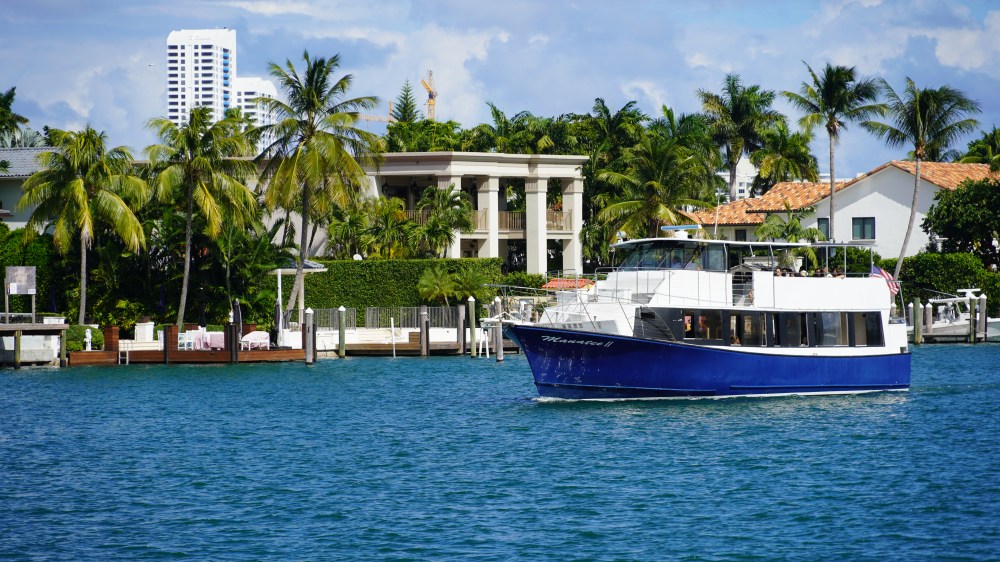 Blue boat on water near luxury homes and palm trees, sunny day.