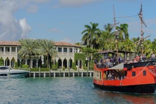 Red pirate ship and yacht in canal with luxury villa backdrop and palm trees.