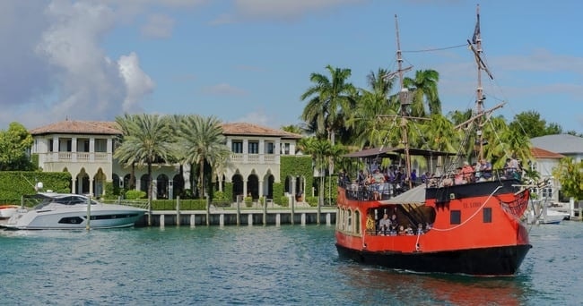 Red pirate ship and yacht in canal with luxury villa backdrop and palm trees.