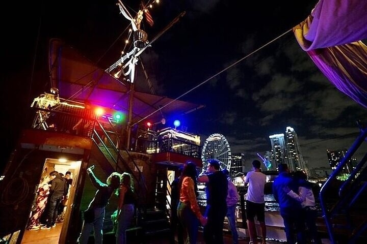 People on a decorated boat at night with city skyline and Ferris wheel in the background.
