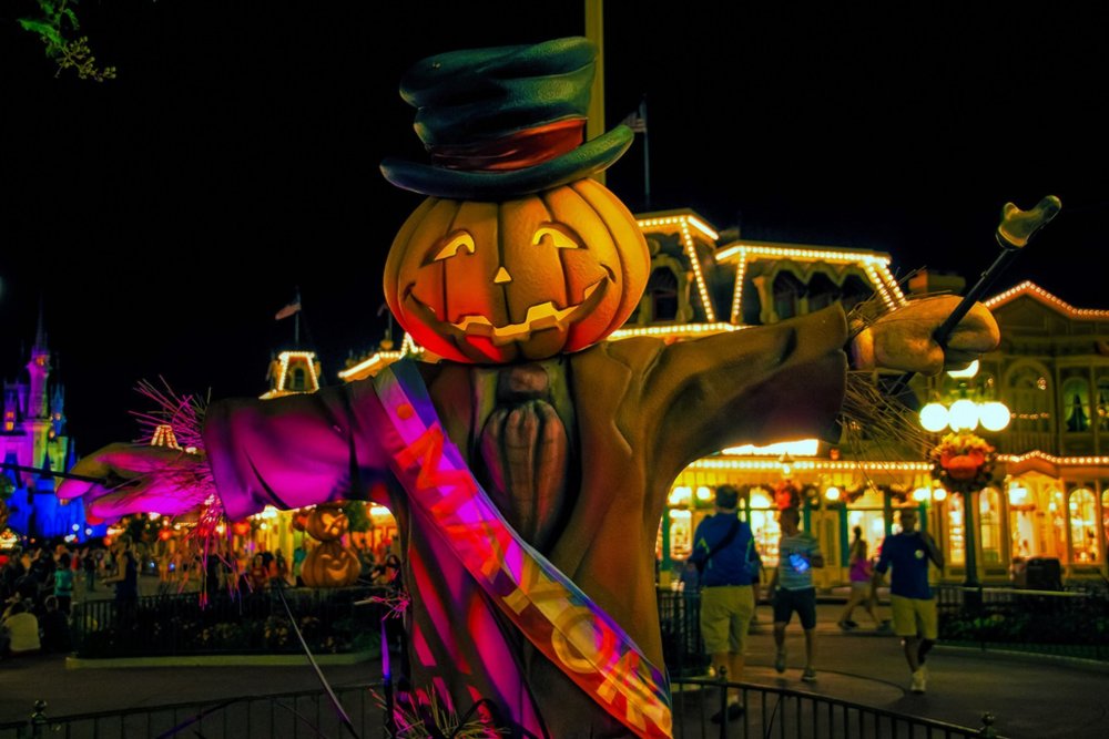 Pumpkin-headed scarecrow dressed as mayor with colorful lighting at a festive nighttime event.
