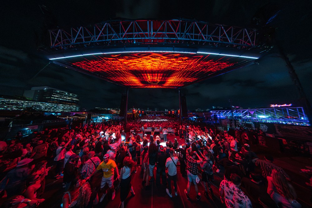 Large outdoor concert at night with vibrant red and blue stage lighting above a crowded audience.