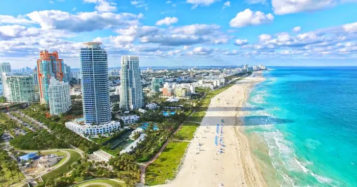 Aerial view of a beach with skyscrapers, turquoise ocean, and blue sky with clouds.