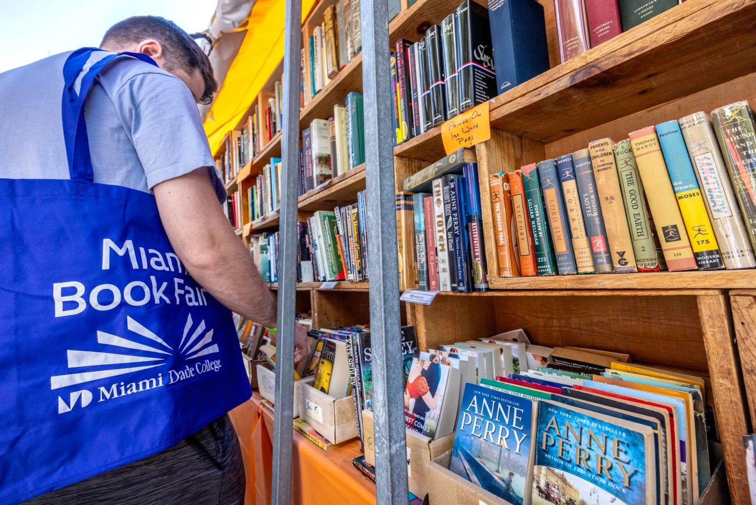 Person browsing bookshelves at Miami Book Fair with a blue tote bag.