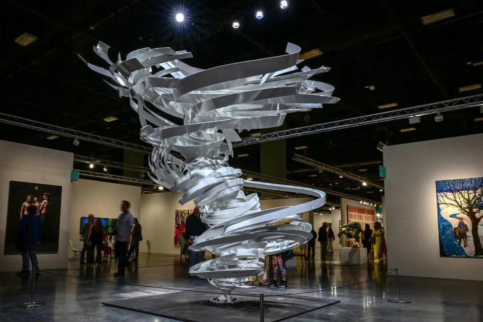 Large twisted metal sculpture in art gallery with people viewing paintings on walls.