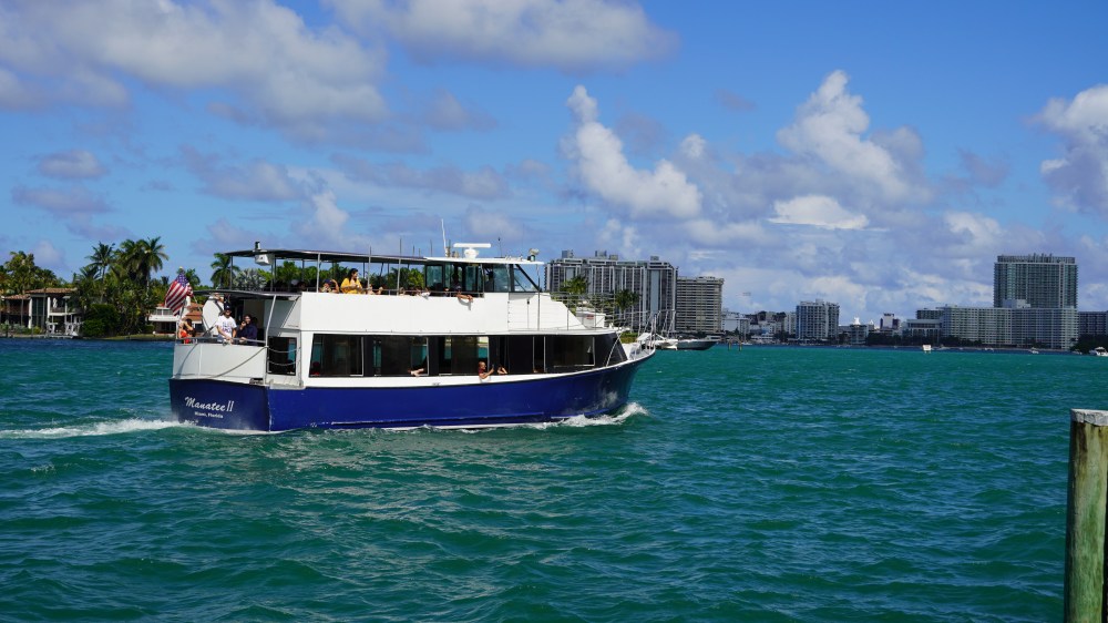 A tour boat on blue water, with city skyline and palm trees in the background.