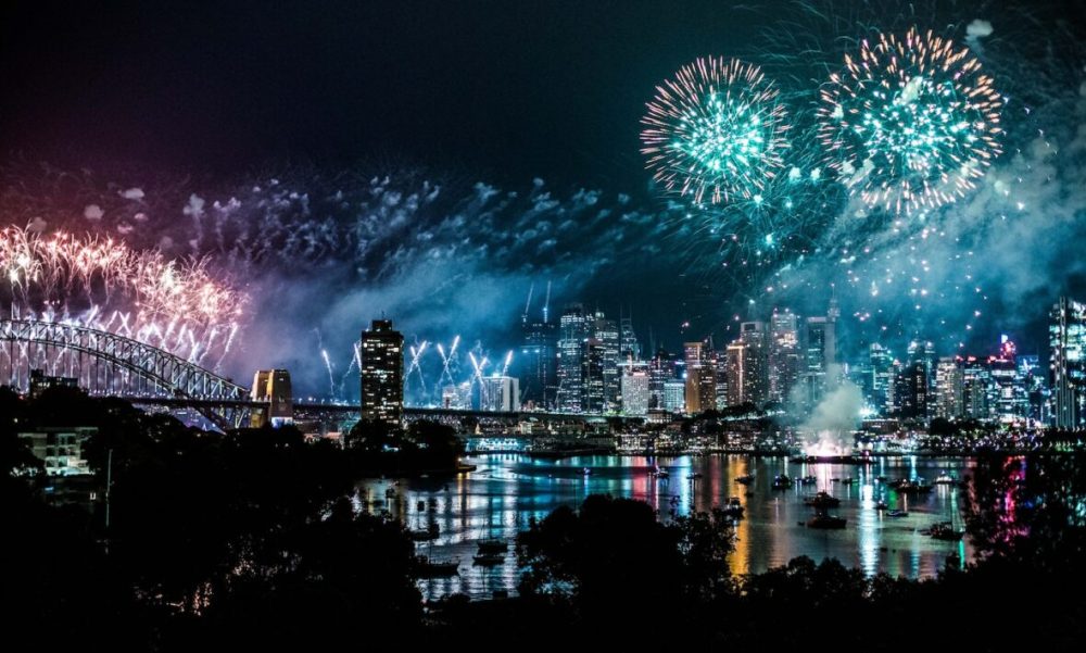 Fireworks over a city skyline at night, reflecting in the water.