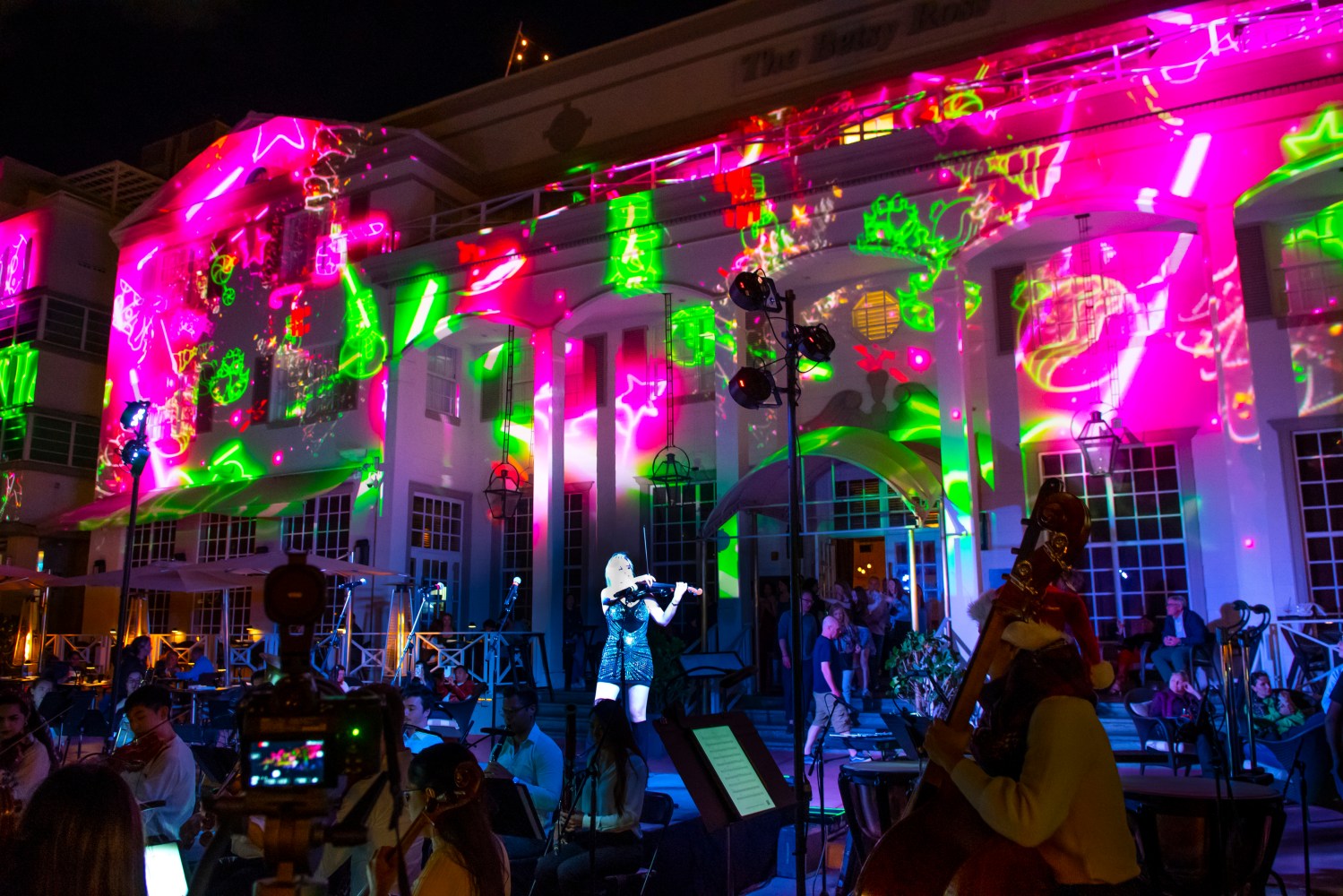 Violinist performs with colorful light projections on a building, surrounded by musicians and audience.