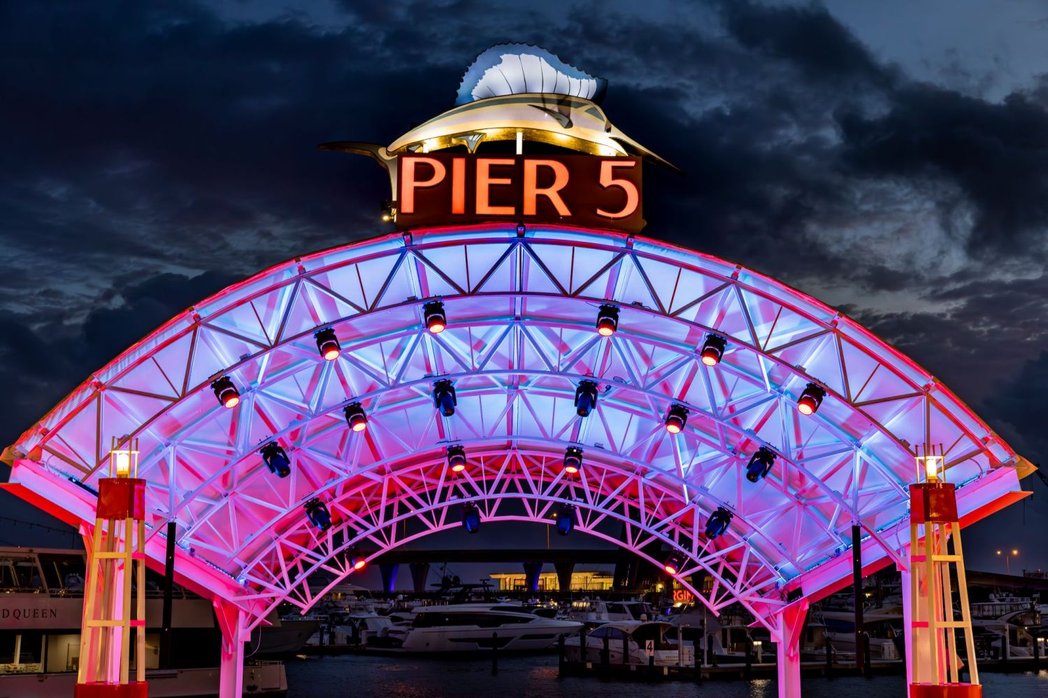 Illuminated Pier 5 sign on an arched metal structure with boats in the background at dusk.