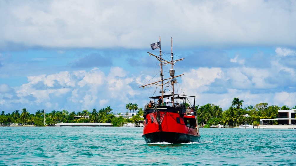Red pirate ship sailing on turquoise water with palm trees and houses in the background.