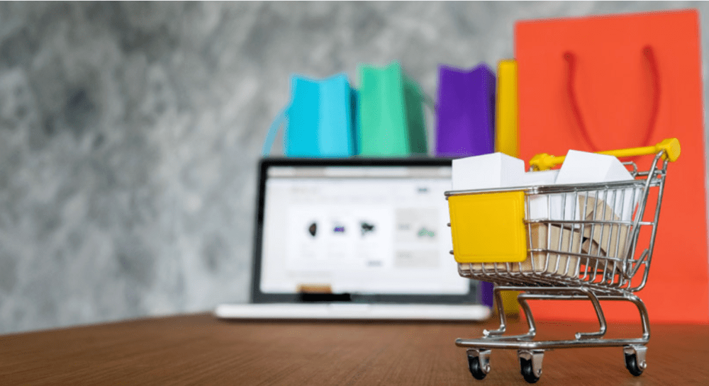 Mini shopping cart with boxes on table, laptop and colored bags in background.