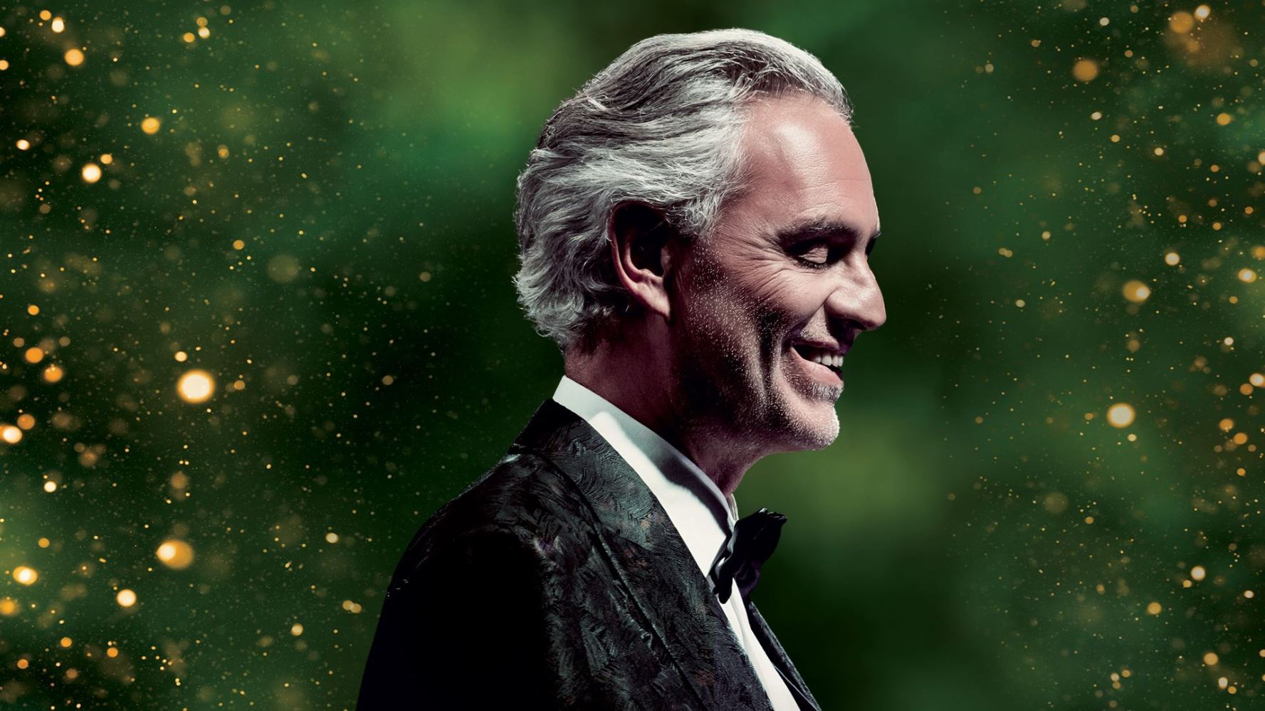 Silver-haired man in black suit smiling, green and gold sparkling background.