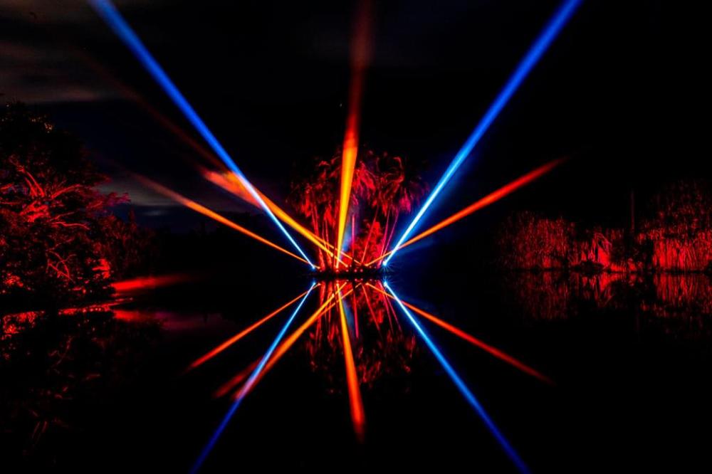 Colorful red and blue laser beams shine over a dark pond with reflections.