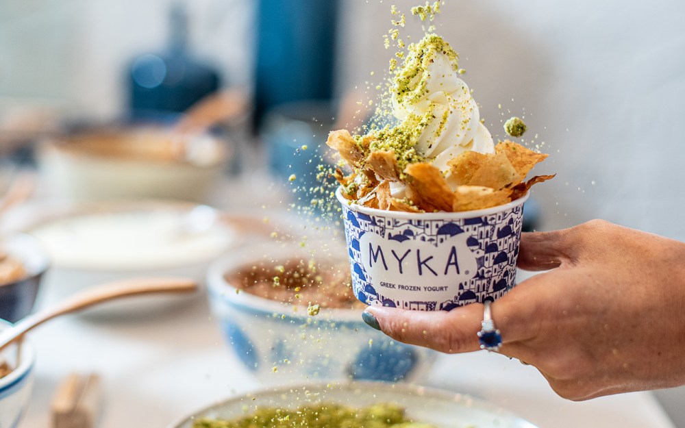 Hand holding Greek frozen yogurt with topping and pistachio sprinkles in a cup labeled 'MYKA'.