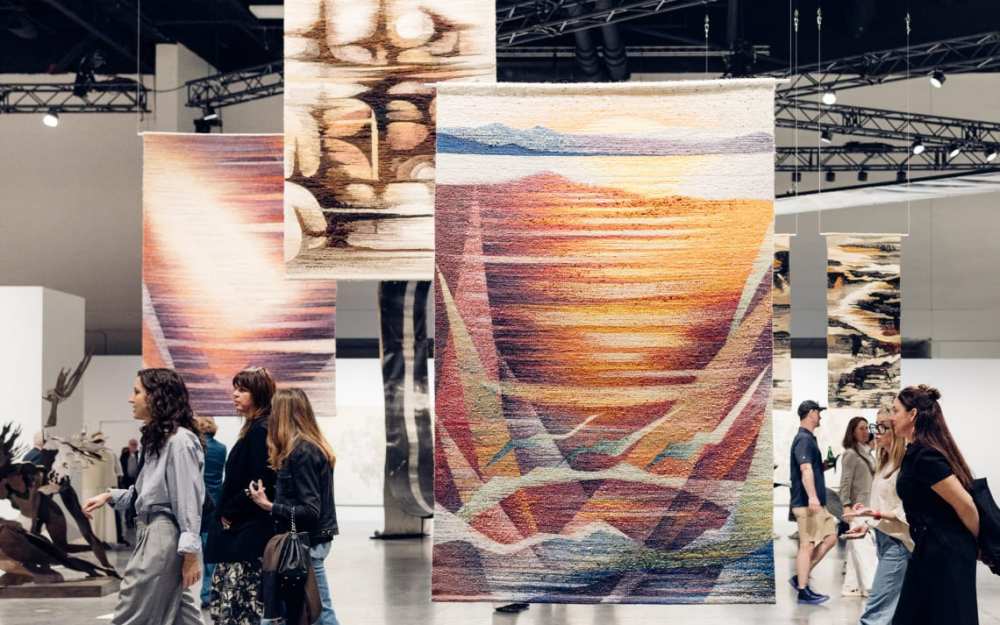 People walking in an art gallery with large, colorful tapestries hanging from the ceiling.