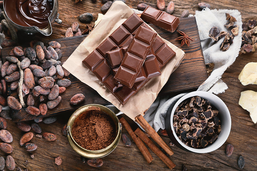 Chocolate bars on wooden board surrounded by cocoa beans, powder, and melted chocolate.