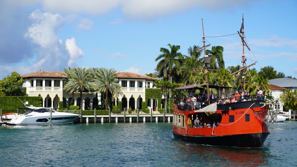Red pirate ship cruising near luxury waterfront mansion with palm trees.