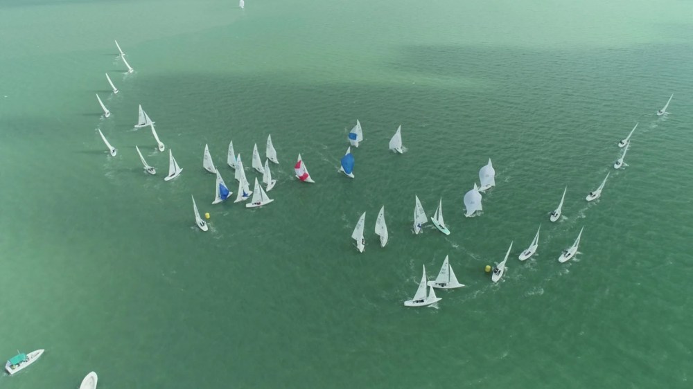 Aerial view of sailboats racing on green water in a semi-circular formation.