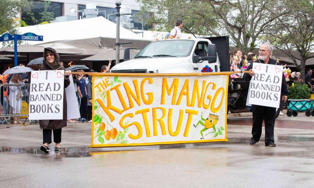 Parade with 'The King Mango Strut' banner and two people holding 'I Read Banned Books' signs.
