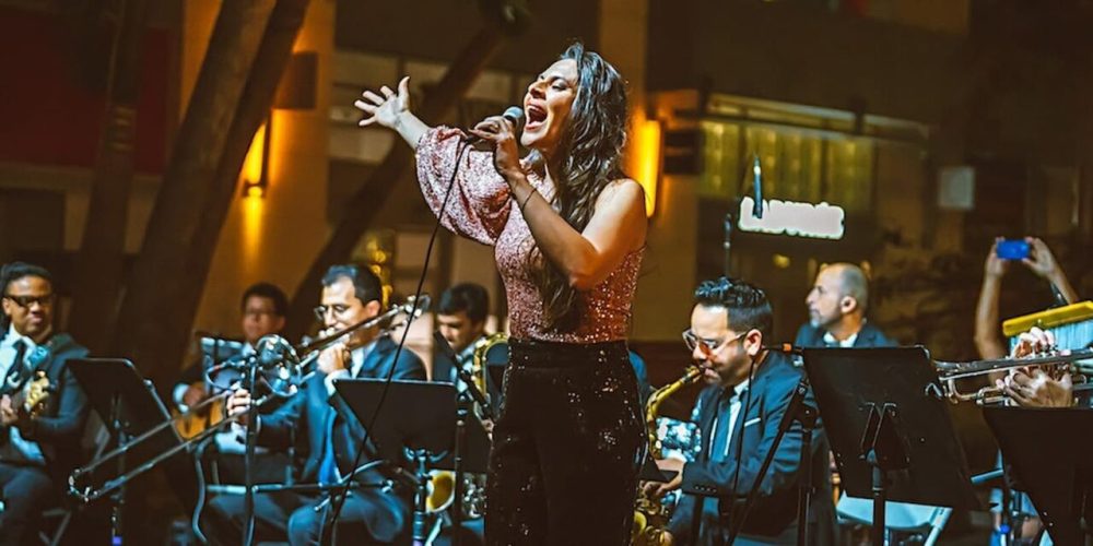 Woman sings passionately on stage, surrounded by a band playing instruments at night.