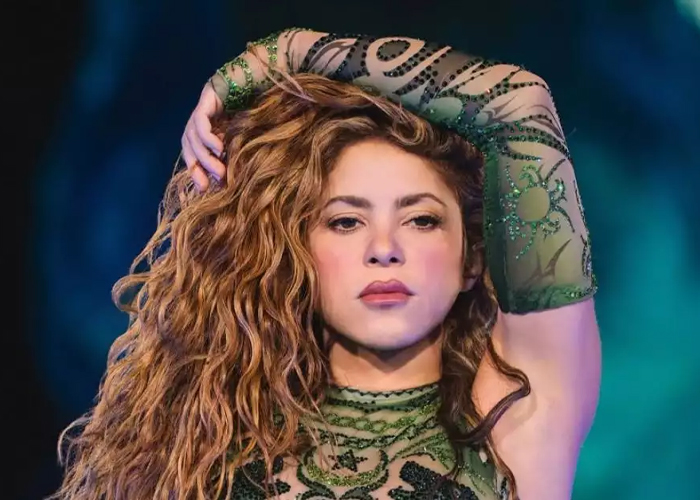 Person with long curly hair poses in a green, patterned outfit with an arm raised above head.