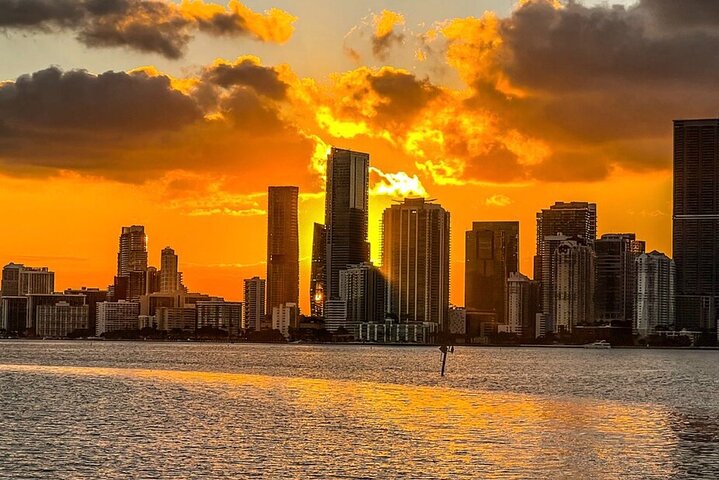 Silhouette of a city skyline at sunset with orange and yellow sky reflecting on water.
