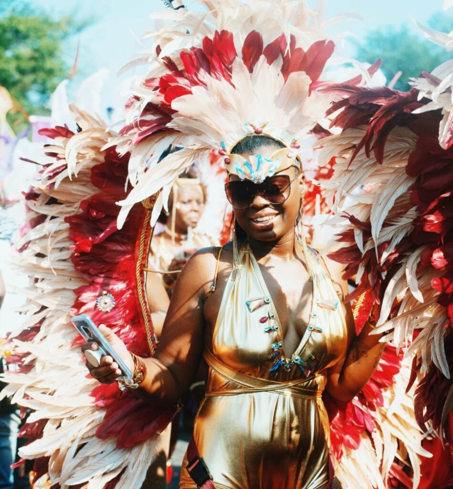 Woman in gold outfit with large feather headdress and wings at a festival, holding a smartphone.