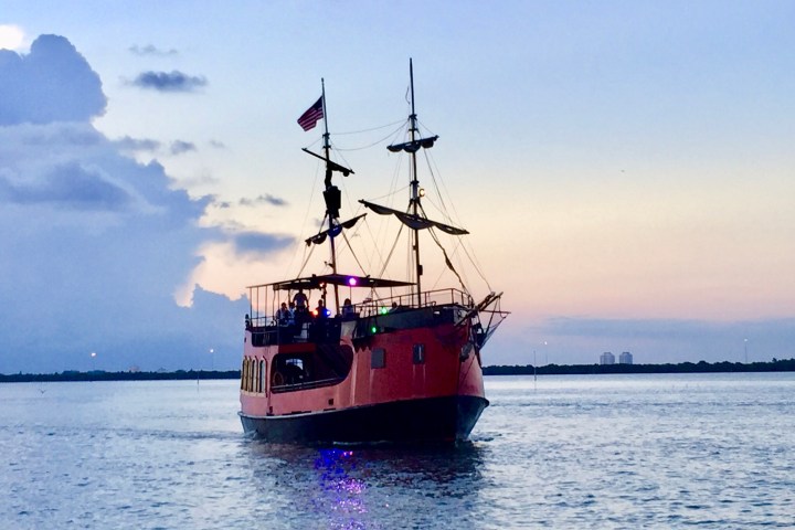 A red ship with sails on calm water at sunset, with people on deck.