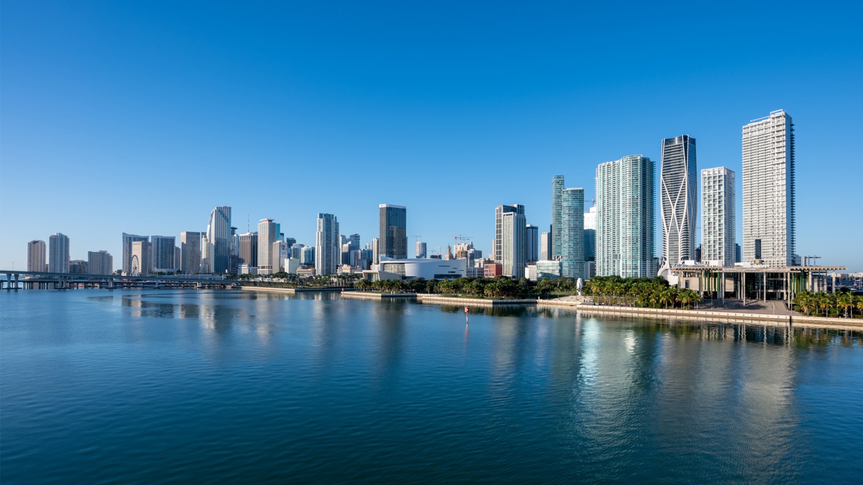 Miami skyline with skyscrapers reflecting on calm water under a clear blue sky.