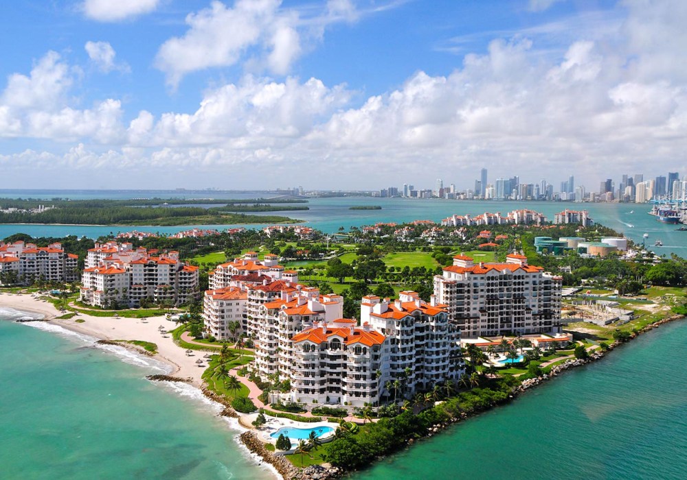 Aerial view of coastal city with white buildings and orange roofs near turquoise water and distant skyline.