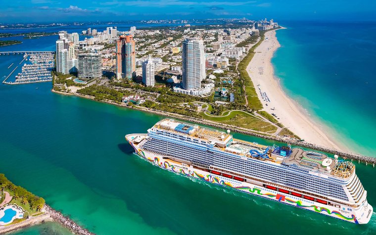 Cruise ship sailing near a coastal cityscape with tall buildings and a sandy beach.