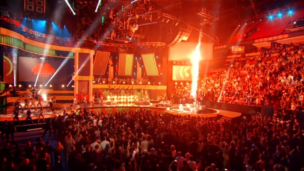 Crowded arena with performers on stage and bright lights, featuring a large flame effect.