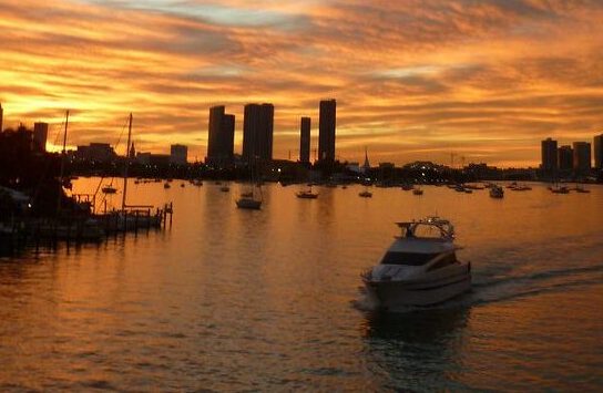 Sunset over city skyline with boats on a calm river.