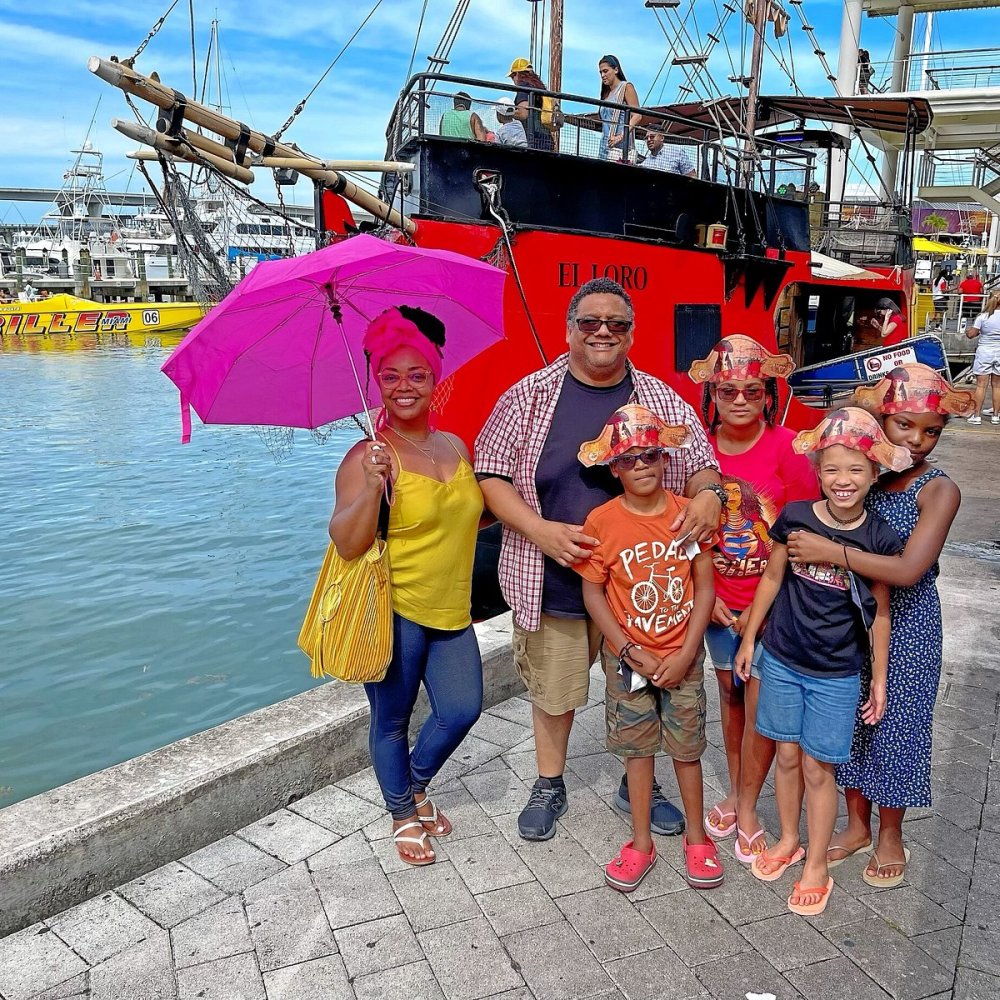 Family posing with pink umbrella and pirate hats by a red boat docked at a harbor.