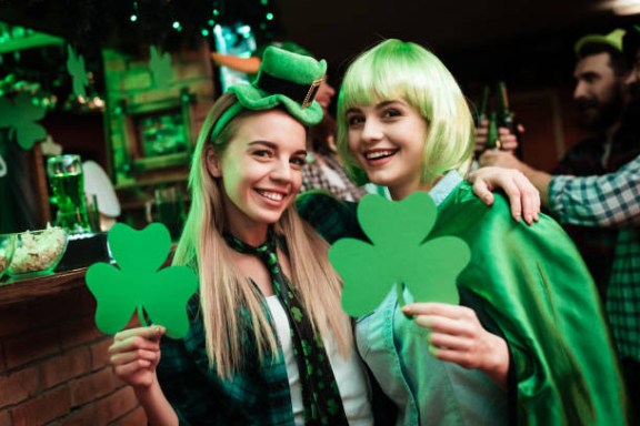 Two women in festive green outfits holding shamrocks, celebrating indoors.