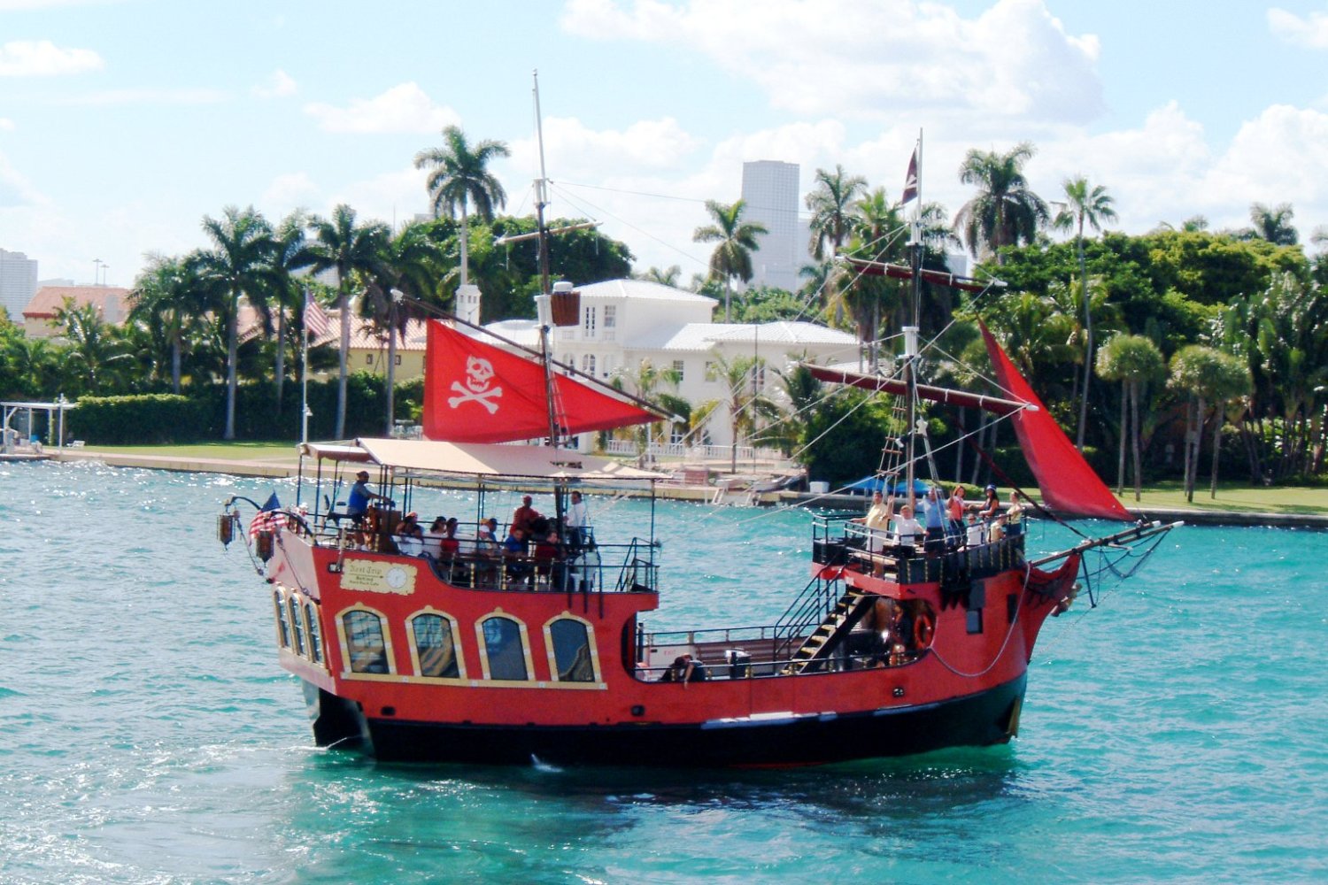 Pirate-themed tour boat with red sails on blue water near palm trees and buildings.