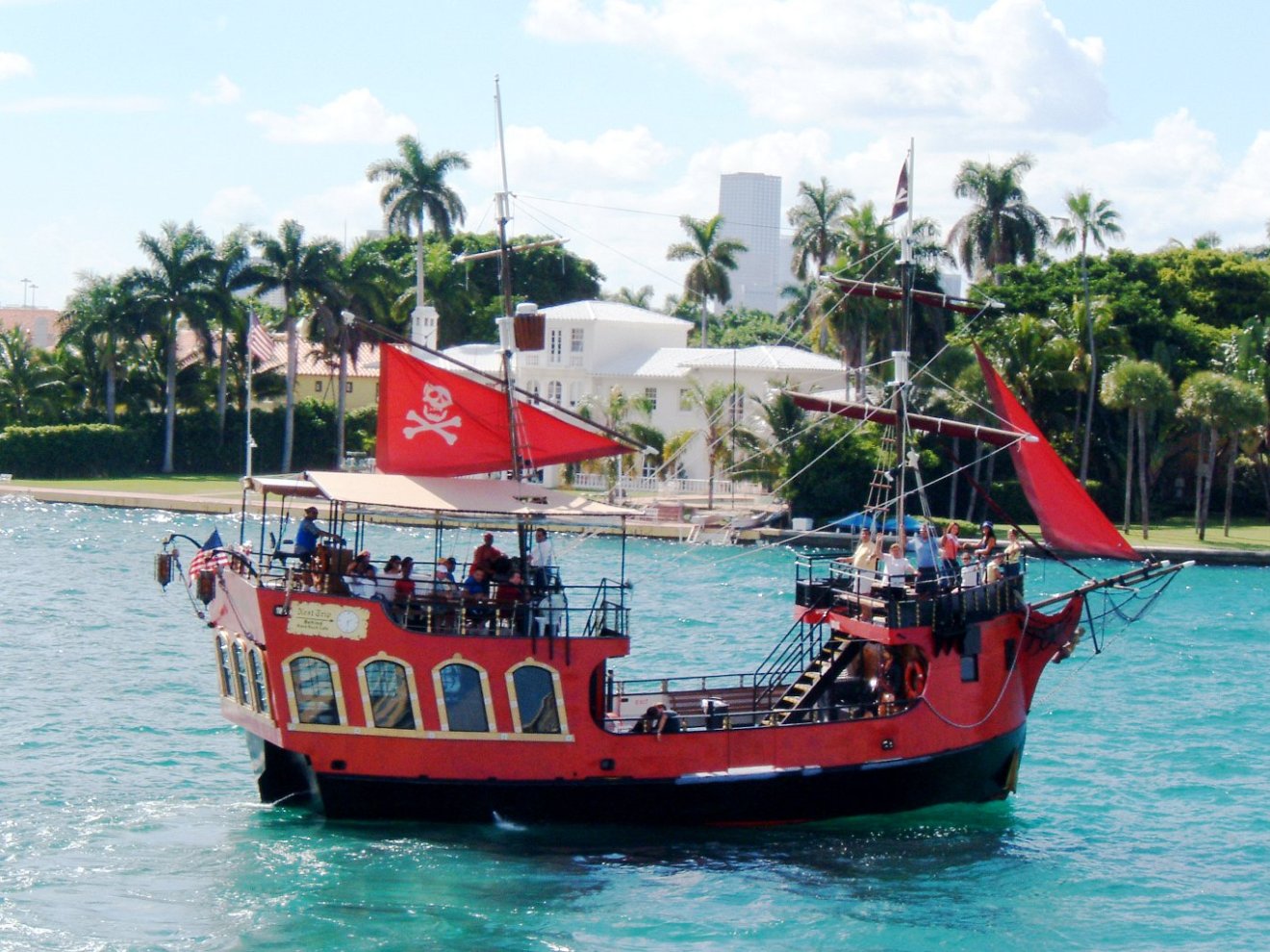 Red pirate-themed ship with people aboard, sailing on a sunny day with palm trees on shore.